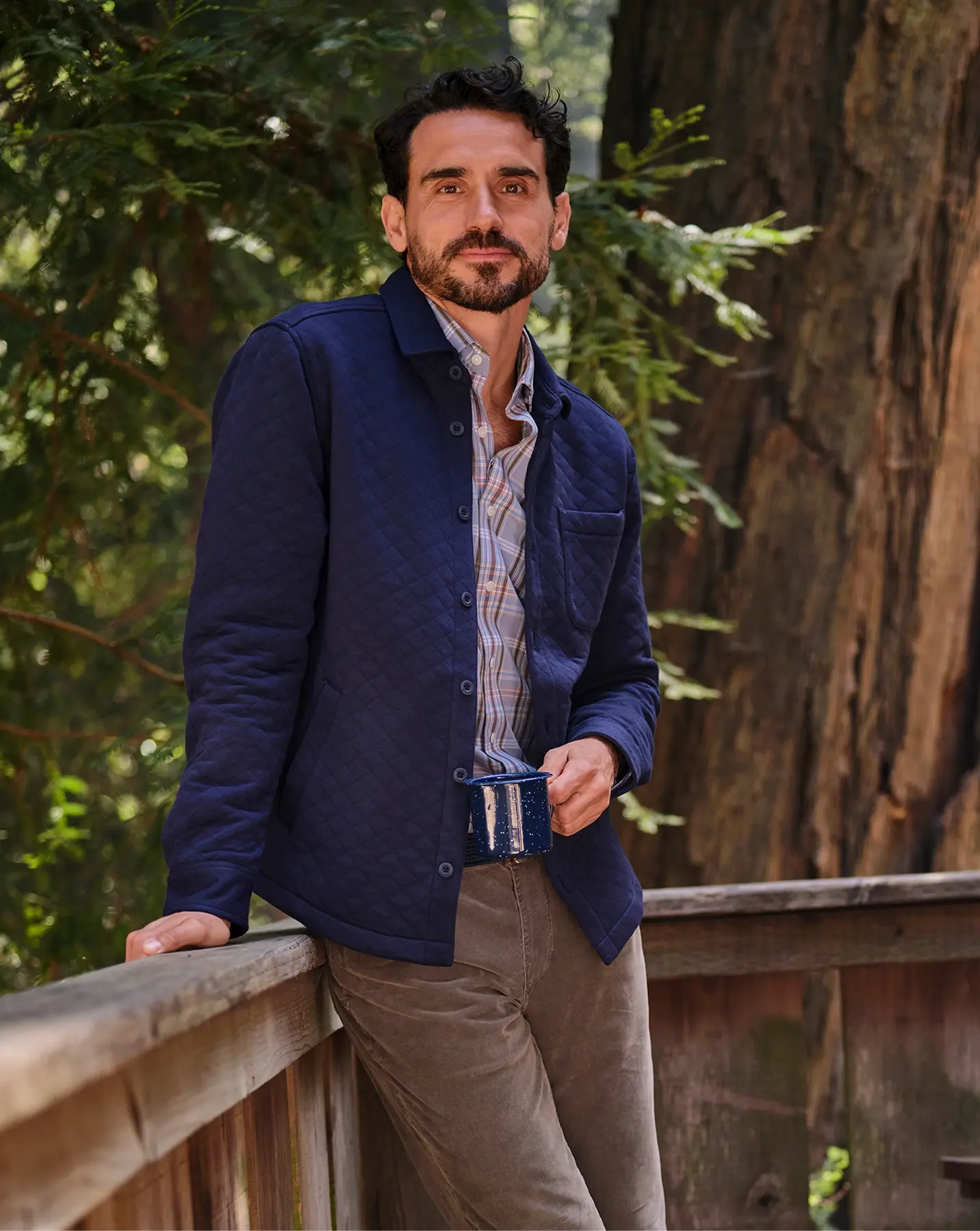 Man in a navy quilted overshirt leaning on a wooden rail under redwoods, holding an enamel mug