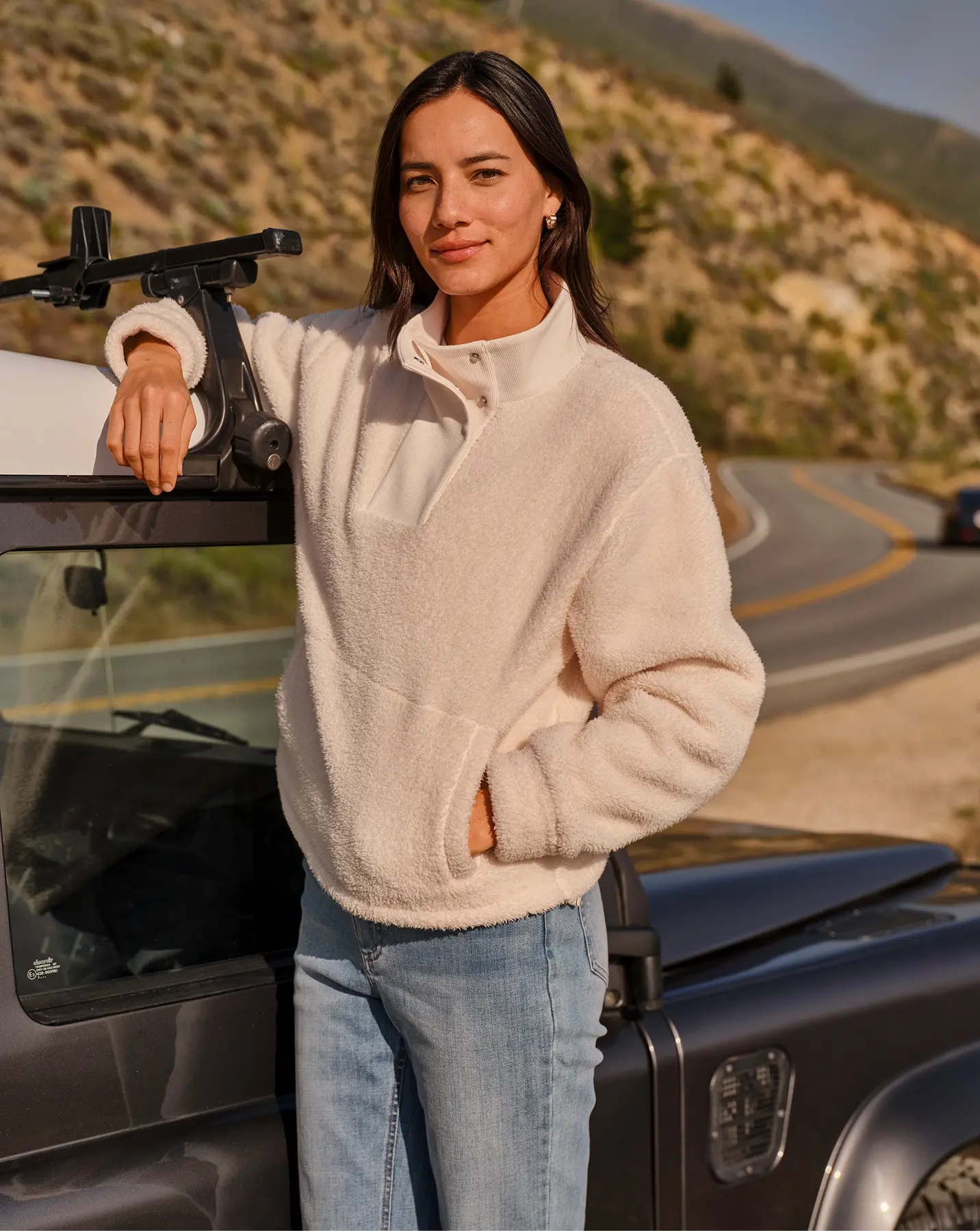 Woman in a cream sherpa pullover and jeans leaning on a dark 4x4 on a coastal road