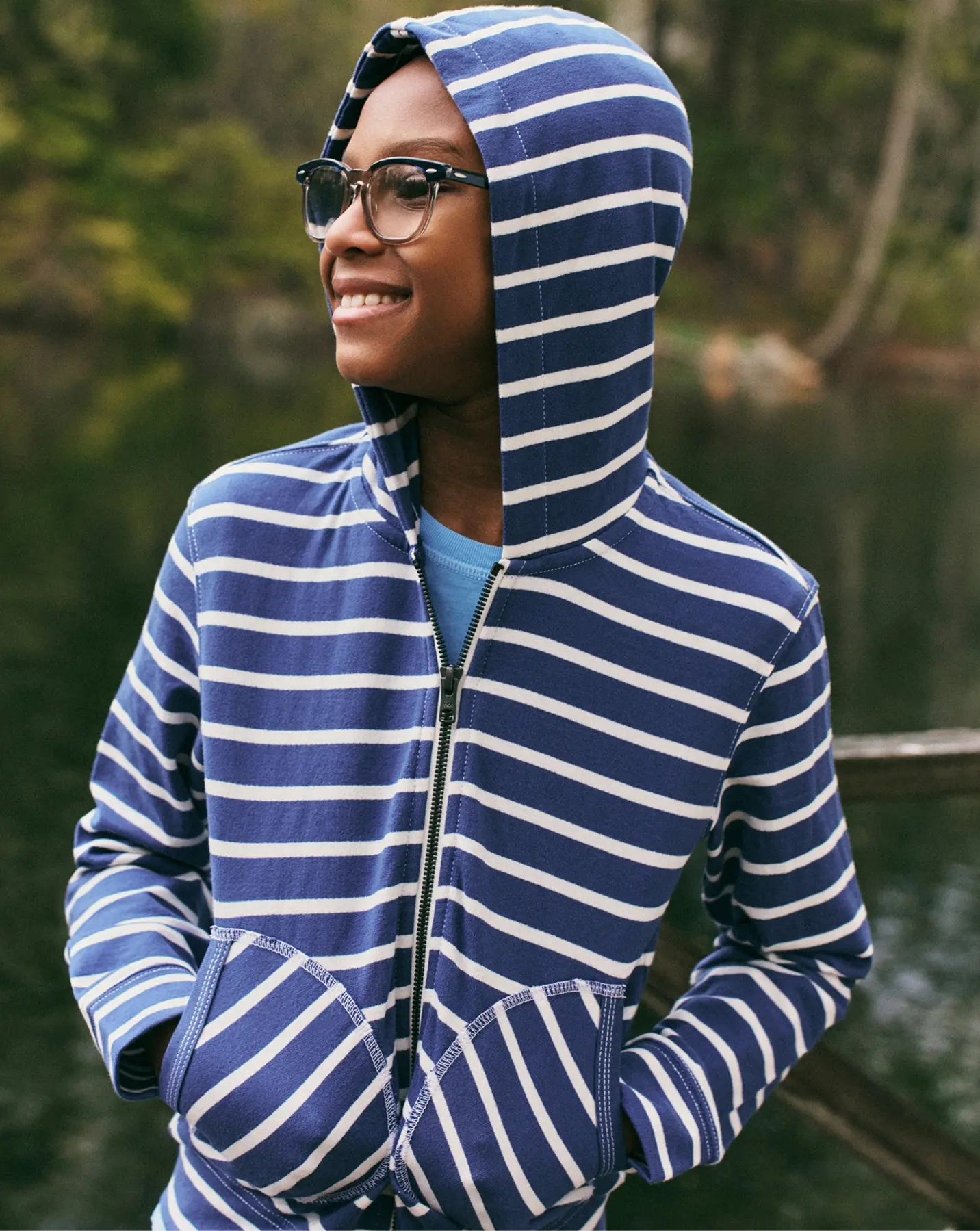 Smiling boy in glasses wearing a blue-and-white striped zip hoodie with the hood up, by a lake