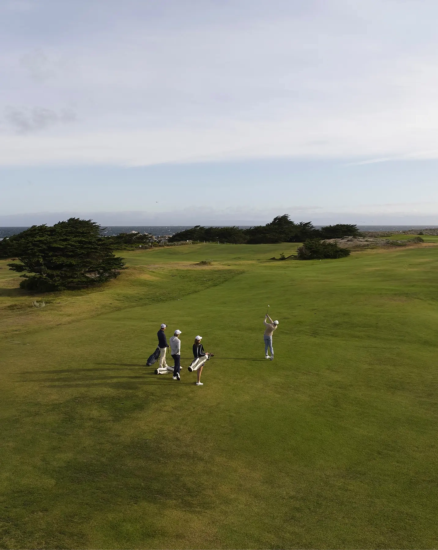 Aerial view of four golfers on a seaside fairway, one mid-swing, cypress trees and ocean behind
