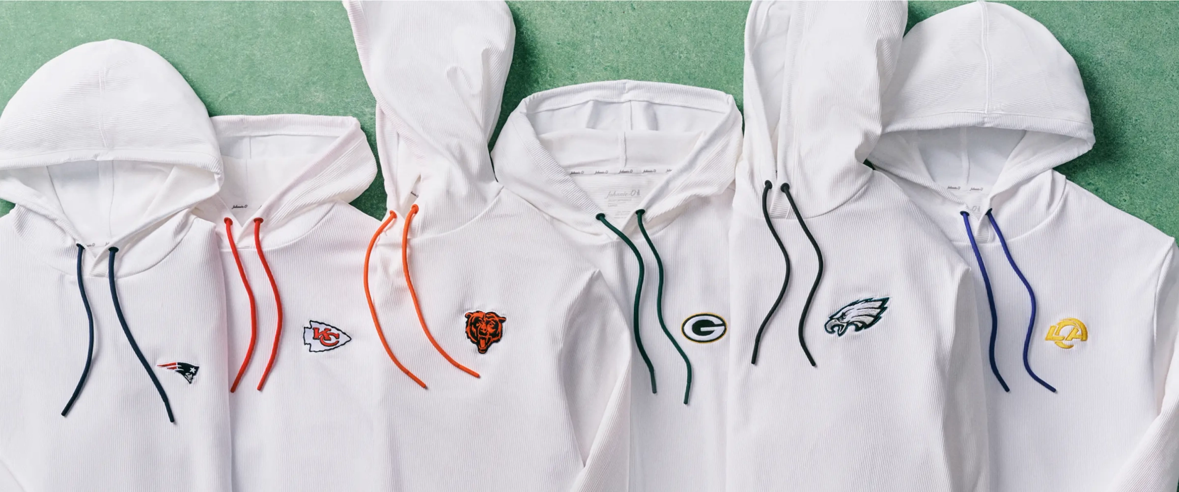 White Johnnie-O hoodies laid on green turf, each embroidered with a pro football team logo and matching drawcords