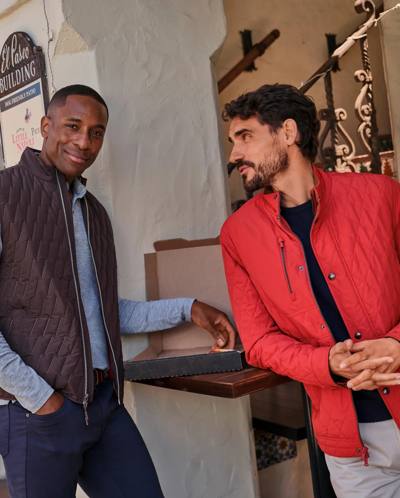Two men leaning on a café ledge, one in a quilted brown vest and one in a red quilted jacket