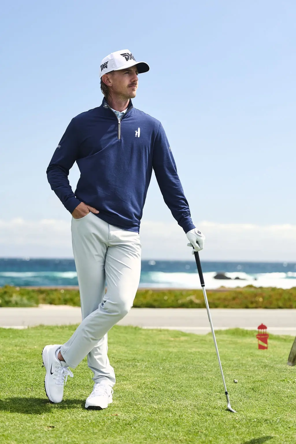 Golfer in a navy Johnnie-O quarter-zip and white cap standing with a club on a tee box by the ocean