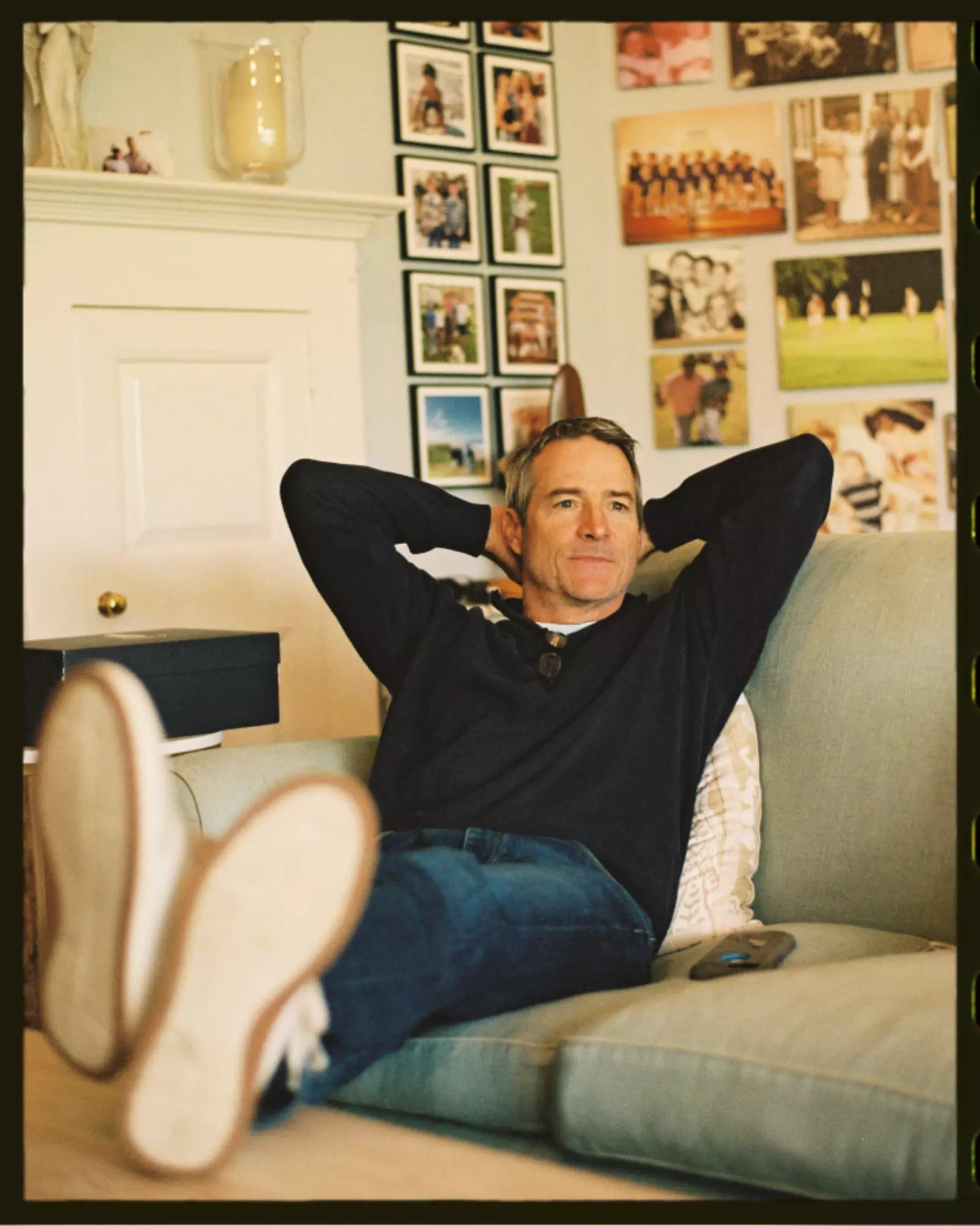 A man in a navy Johnnie-O polo leaning back on a sofa with his hands behind his head, a wall of family photos behind him