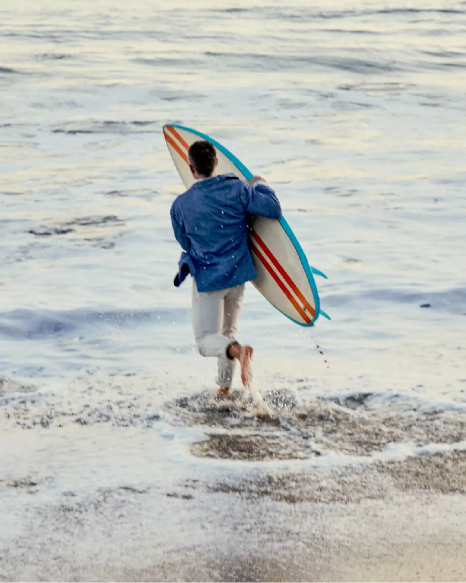 A surfer in a navy Johnnie-O pullover walking into the shallows with a striped board under his arm