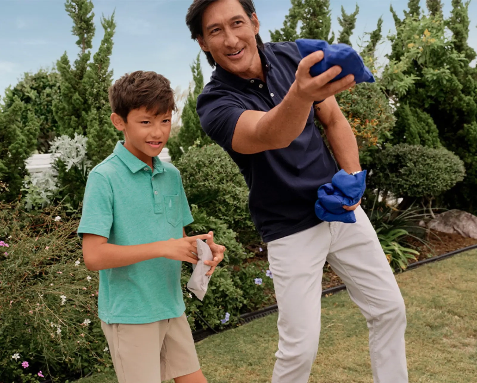 A father in a navy polo tossing a beanbag beside his son in a green polo, hedges and a garden behind them